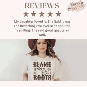 Op de afbeelding: Een cr&egrave;mekleurig T-shirt met korte mouwen en bruine tekst met de tekst BLAME IT ON ALL MY ROOTS, met een grafische omtrek van de staat Michigan in het midden. Het shirt wordt gedragen door een persoon met een bruine hoed met brede rand.