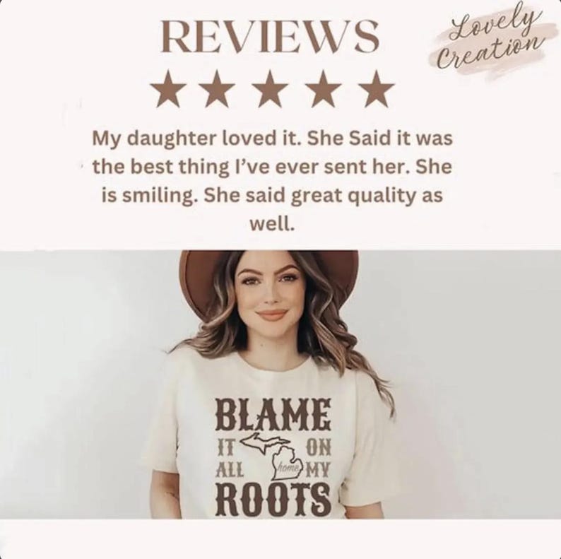 Op de afbeelding: Een cr&egrave;mekleurig T-shirt met korte mouwen en bruine tekst met de tekst BLAME IT ON ALL MY ROOTS, met een grafische omtrek van de staat Michigan in het midden. Het shirt wordt gedragen door een persoon met een bruine hoed met brede rand.