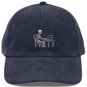May include: A navy blue corduroy baseball cap featuring a white embroidered graphic of a skeleton sitting at a desk and working on a laptop. The laptop screen displays the code symbol consisting of a forward slash between two angle brackets.