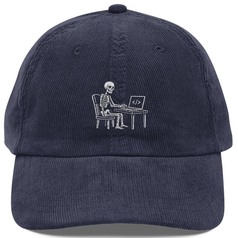 May include: A navy blue corduroy baseball cap featuring a white embroidered graphic of a skeleton sitting at a desk and working on a laptop. The laptop screen displays the code symbol consisting of a forward slash between two angle brackets.