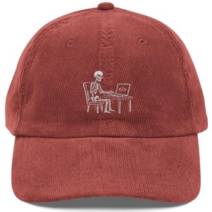 May include: A brick red corduroy baseball cap featuring a white embroidered graphic of a skeleton sitting at a desk and working on a laptop. The laptop screen displays a code symbol consisting of a slash between two angle brackets.