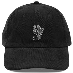 Puede incluir: Una gorra de béisbol de pana negra con un gráfico bordado en blanco de un esqueleto tocando el arpa. La gorra tiene una corona estructurada, una visera curva y un botón en la parte superior. El diseño está centrado en el panel frontal.