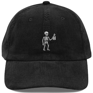 May include: A black corduroy baseball cap featuring an embroidered white graphic of a skeleton holding a steaming mug. The cap has a structured crown, a curved brim, and a button at the top centre. The embroidery is positioned centrally on the front panel.