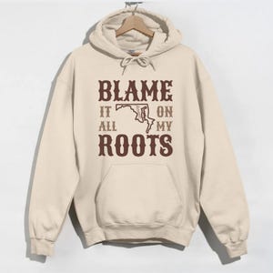 May include: A beige hoodie with a brown graphic print that says "Blame it all on my roots" with an outline of the state of Maryland.
