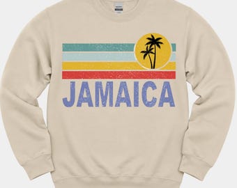May include: A beige crewneck sweatshirt featuring a distressed graphic of the word JAMAICA in blue letters below horizontal stripes in teal, light blue, yellow, and red, with a yellow circle containing a black silhouette of two palm trees.