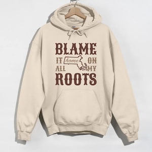 Puede incluir: Una sudadera con capucha beige con un gráfico marrón que dice "Blame it home on all my roots" con un contorno del estado de Massachusetts.