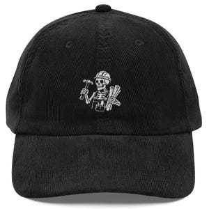 Peut inclure: Une casquette de baseball en velours côtelé noir avec une broderie blanche représentant un squelette portant un casque de chantier et une salopette. Le squelette tient un marteau et des plans enroulés, avec une pochette à outils à la taille.