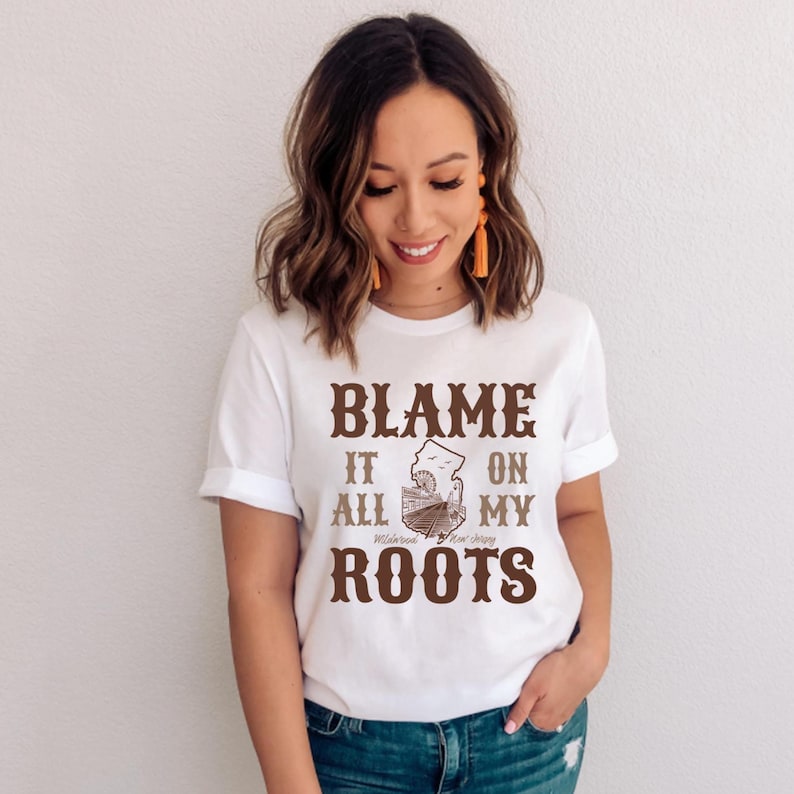 Op de afbeelding: Een wit T-shirt met korte mouwen en bruine tekst die luidt BLAME IT ON ALL MY ROOTS. De afbeelding bevat een kleine illustratie van een reuzenrad en treinsporen, met de tekst Wildwood New Jersey onder de afbeelding gedrukt.