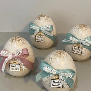 Peut inclure: Quatre œufs de Pâques blancs décorés de paillettes dorées et noués de rubans bleu pastel et rose. Chaque œuf a un charme doré avec le texte "Kalo Pascha" en grec.