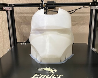 May include: A white 3D printed plastic helmet with a design resembling a superhero mask. The helmet is sitting on a black platform with the word "Ender" printed on it.