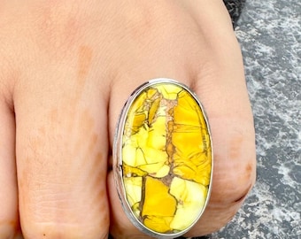 May include: A silver ring featuring a large, oval yellow gemstone. The stone displays a distinctive pattern of yellow and brown shades. The ring is worn on a hand adorned with henna and a dark green and red bracelet.