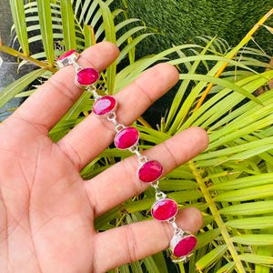 Pode incluir: Uma pulseira de prata com pedras preciosas vermelhas ovais e vibrantes. As pedras são ligadas com conectores de prata. A pulseira é exibida em um fundo de folhagem verde.
