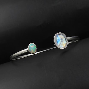 May include: A silver bracelet featuring two oval-shaped gemstones. The larger gemstone is iridescent and the smaller gemstone is a pale green opal.
