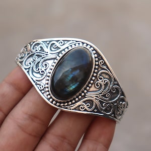 May include: Silver-toned cuff bracelet featuring an oval, iridescent gemstone. The bracelet has an ornate, swirling design around the stone and along the band. The gemstone displays shades of blue, green, and teal.