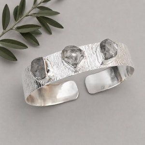 Peut inclure: Un bracelet manchette en argent avec une surface métallique brossée et texturée, orné de trois pierres brutes translucides gris-blanc serties dans des chatons en argent poli. Le bracelet présente une ouverture à l'arrière pour l'ajustement.