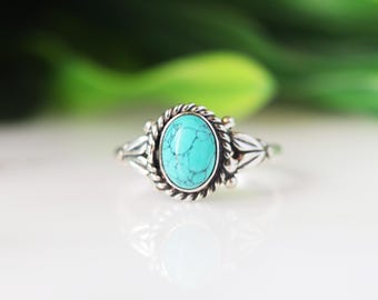 May include: A silver ring with an oval turquoise stone. The stone is set in a detailed silver setting with a rope-like border. The ring has decorative silver accents on the sides, creating a vintage or bohemian style.