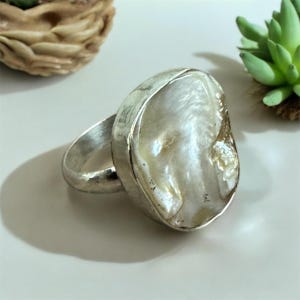 May include: A silver ring with a large, white, pearlescent stone set in a bezel setting. The stone has a natural, organic shape.