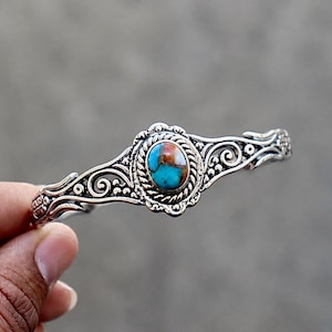 Könnte beinhalten: Ein silberfarbenes Armband mit einem ovalen Türkisstein mit braunen Matrixmustern in der Mitte. Das Metallband ist mit komplizierten Schnörkeln, Perlenakzenten und einer gedrehten Kordelkante verziert, die den Stein umgibt.