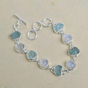 Puede incluir: Un brazalete de plata con piedras preciosas de aguamarina y piedra lunar alternas. El brazalete tiene un cierre de barra en T.