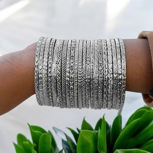 May include: A collection of silver bangles with various etched designs, stacked on a wrist. The bangles feature patterns like floral, geometric, and textured lines. The image is set against a backdrop of green foliage.