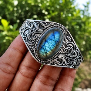 Può includere: Un bracciale a fascia in argento con una pietra preziosa di labradorite ovale con iridescenza blu al centro. La fascia in metallo è decorata con intricati motivi a volute in rilievo e un bordo a corda intrecciata attorno alla pietra.