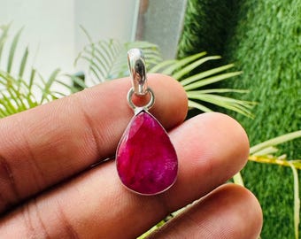 May include: A teardrop-shaped pendant with a vibrant red gemstone. The gemstone is set in a silver-colored metal setting with a loop for a chain. The pendant is held in a hand against a background of green foliage.