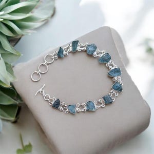 May include: A silver bracelet with a double chain design features light blue aquamarine stones. The bracelet has a toggle clasp.