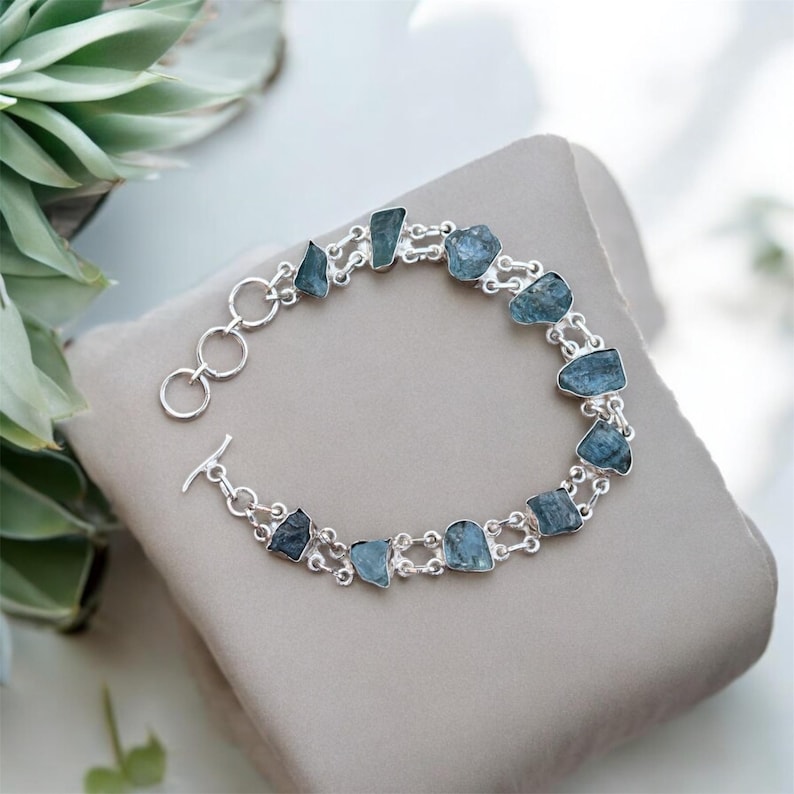 May include: A silver bracelet with a double chain design features light blue aquamarine stones. The bracelet has a toggle clasp.