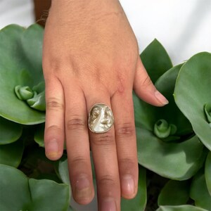May include: A silver ring with a white, face-shaped stone. The stone has a carved design of a face.