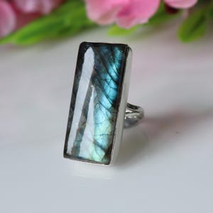 May include: A silver ring with a rectangular labradorite stone. The stone has iridescent blue, green, and grey tones. The ring's setting is a simple, polished silver band.