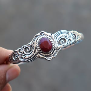 Può includere: Braccialetto in argento con un design elaborato e una pietra preziosa rossa al centro.