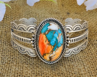 May include: A silver cuff bracelet featuring a large oval center stone with a marbled pattern of bright orange, turquoise blue, and white. The metal band has a scalloped edge with stamped detailing and a twisted rope border around the stone.