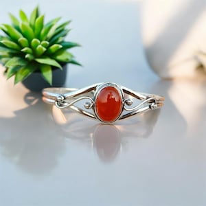 May include: A silver bangle bracelet with an orange carnelian gemstone. The bracelet has a delicate, ornate design.