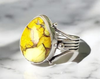 May include: A silver ring with a large, teardrop-shaped yellow stone with brown veining. The stone is set in a silver bezel and the ring has a multi-band design on the band. The ring is displayed on a white marble surface.
