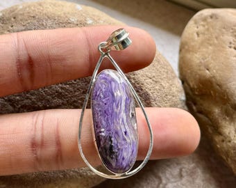 Peut inclure: Un pendentif en forme de goutte d'eau avec une pierre de charoïte ovale polie. La pierre présente des teintes violettes avec des veines blanches et noires, sertie dans un cadre en métal argenté. Le pendentif est conçu pour être porté en collier.