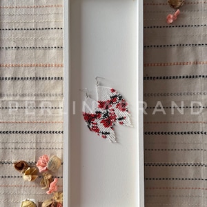 Peut inclure: Une paire de boucles d'oreilles en perles blanches, rouges et noires avec un motif géométrique. Les boucles d'oreilles sont présentées dans un plateau rectangulaire blanc.