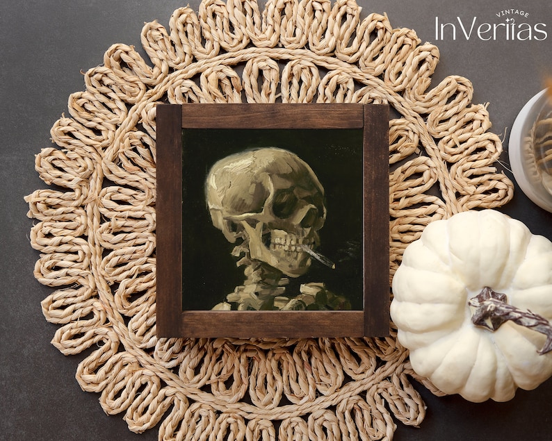 May include: A framed painting of a human skull with a cigarette in its mouth. The skull is painted in a realistic style with a dark background. The frame is dark brown wood.