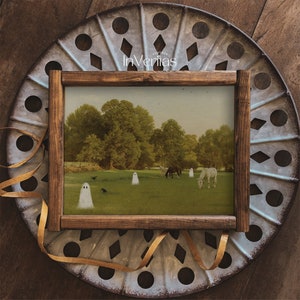 May include: A framed print featuring a pastoral scene with horses grazing in a field, with ghostly figures and crows. The wooden frame surrounds the artwork, set against a decorative metal background. The text "Vintage InVeritas" is visible.