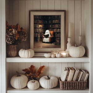 Pode incluir: Uma aconchegante exibi&ccedil;&atilde;o de prateleira com tema de Halloween. Apresenta uma pintura emoldurada de um fantasma lendo em uma biblioteca, ab&oacute;boras brancas de v&aacute;rios tamanhos, velas, folhas secas e uma cesta de livros. A paleta de cores &eacute; neutra com toques quentes.