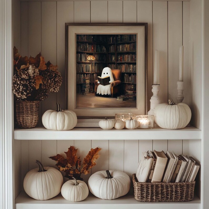 Pode incluir: Uma aconchegante exibi&ccedil;&atilde;o de prateleira com tema de Halloween. Apresenta uma pintura emoldurada de um fantasma lendo em uma biblioteca, ab&oacute;boras brancas de v&aacute;rios tamanhos, velas, folhas secas e uma cesta de livros. A paleta de cores &eacute; neutra com toques quentes.
