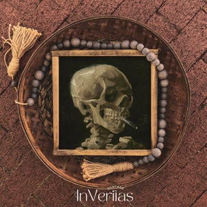 May include: A framed painting of a human skull with a cigarette in its mouth. The painting is set against a dark background and is surrounded by a wooden frame. The painting is displayed on a woven tray with a beaded garland.