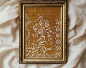 Puede incluir: Una obra de arte enmarcada con un marco dorado y un fondo marrón. La obra presenta un diseño floral blanco con detalles intrincados.