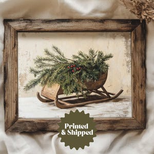 May include: A framed print depicting a wooden sled laden with evergreen boughs and red berries. The artwork has a vintage feel, with a distressed wooden frame and a neutral backdrop. The text "Printed & Shipped" is visible.