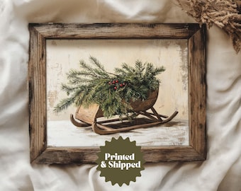May include: A framed print depicting a wooden sled laden with evergreen boughs and red berries. The artwork has a vintage feel, with a distressed wooden frame and a neutral backdrop. The text "Printed & Shipped" is visible.
