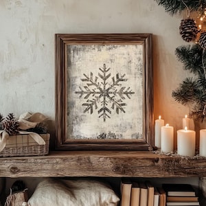 Puede incluir: Una impresión enmarcada de un copo de nieve negro sobre un fondo blanco con una textura desgastada. El marco es de madera marrón y la impresión está sobre un estante de madera con velas y libros.