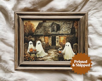 May include: A framed print shows three ghost figures at a table outside a cottage. The ghosts are draped in white sheets, enjoying tea. The artwork has a warm, autumnal colour palette, set in a brown wooden frame. The image is a charming Halloween scene.