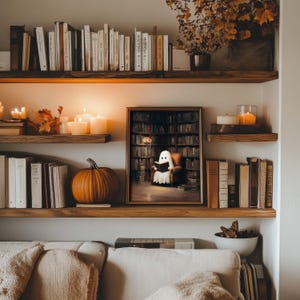 Pode incluir: Uma cena acolhedora de sala de estar com prateleiras de madeira cheias de livros, velas e uma ab&oacute;bora. Uma obra de arte emoldurada retrata um fantasma lendo em uma biblioteca. O quarto &eacute; decorado em tons quentes de outono.