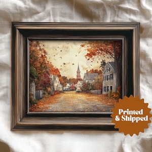 May include: Framed print of a quaint autumn town scene. The artwork depicts a street lined with houses and trees in fall colors. The frame is a dark, rustic wood. The print has a warm, inviting aesthetic.