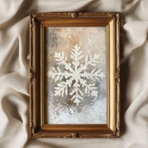 Puede incluir: Una pintura enmarcada de un copo de nieve blanco sobre un fondo beige y marrón. El marco es dorado.