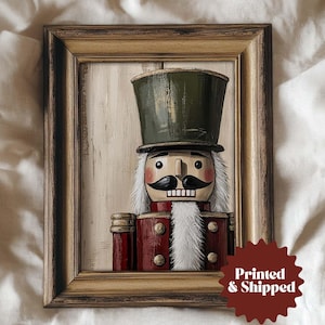 May include: Framed artwork featuring a painted nutcracker soldier. The nutcracker has a green top hat, a black mustache, and a red coat. The artwork is in a wooden frame with a distressed finish. The text "Printed & Shipped" is on the bottom right.